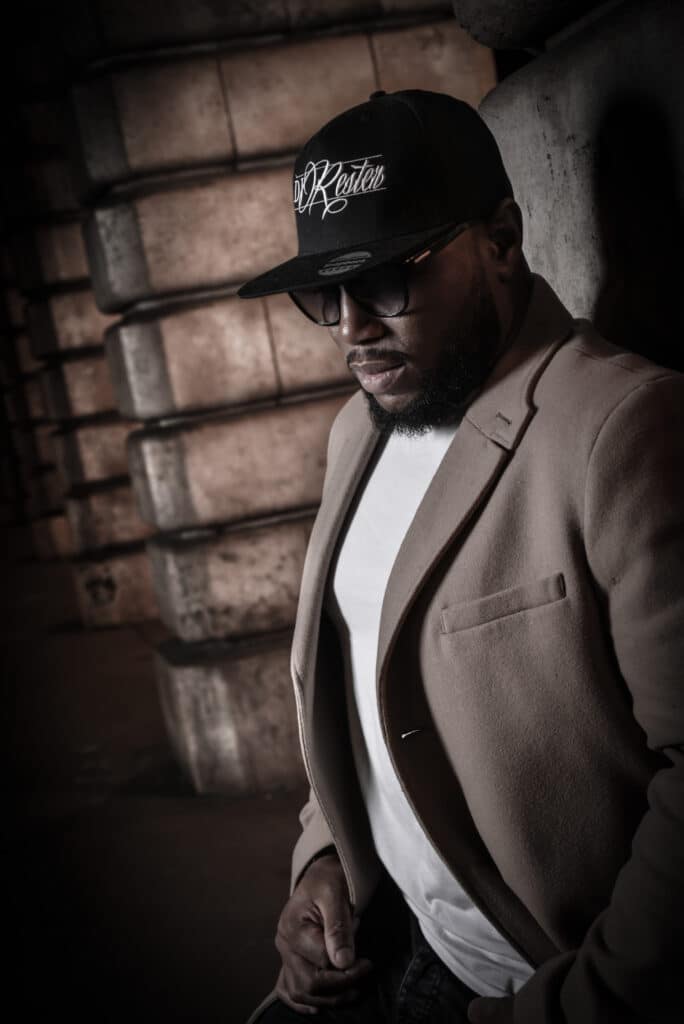 Portrait DJ Rosten: Style urbain en veste et casquette Homme en veste beige et casquette noire DJ Rosten, lunettes, penché contre mur sombre, regardant vers le bas près de piles.