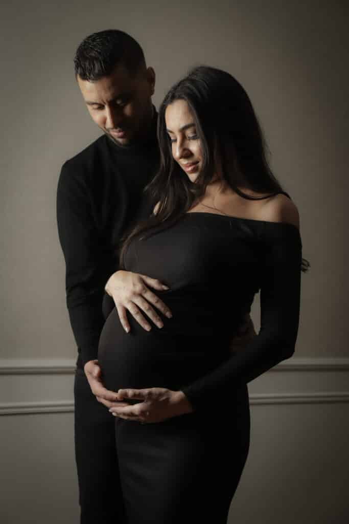 Séance photo grossesse couple | Maternité tendre Portrait intime d'un couple enceinte. L'homme serre la femme par derrière, leurs mains sur son ventre rond. Tendresse et attente.