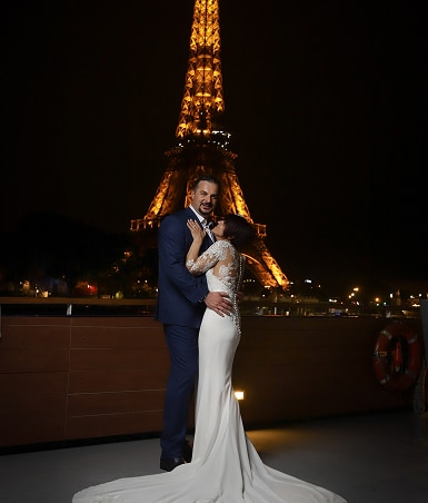 Couple élégant s'embrassant devant la Tour Eiffel illuminée à Paris la nuit. Robe de mariée blanche, costume bleu marine. Ambiance romantique sur la Seine.