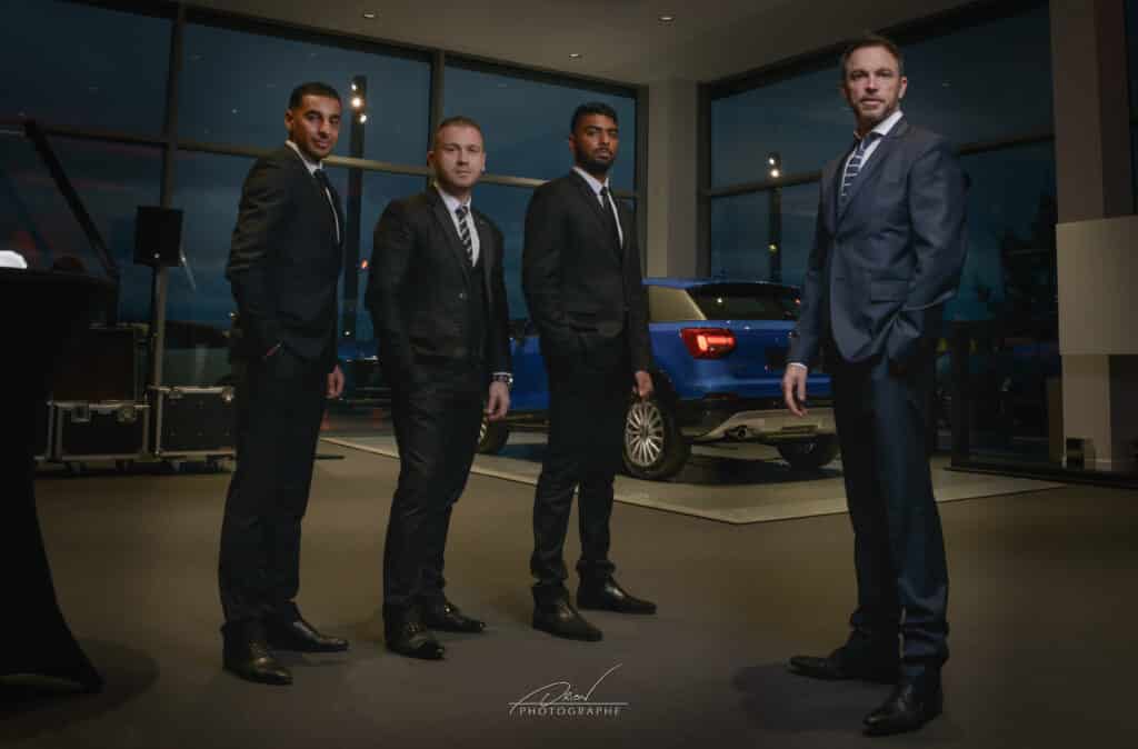 Quatre hommes en costume posent dans un showroom moderne avec une voiture bleue en arrière-plan. Équipement d'événement visible. Watermark Lion Photographe.