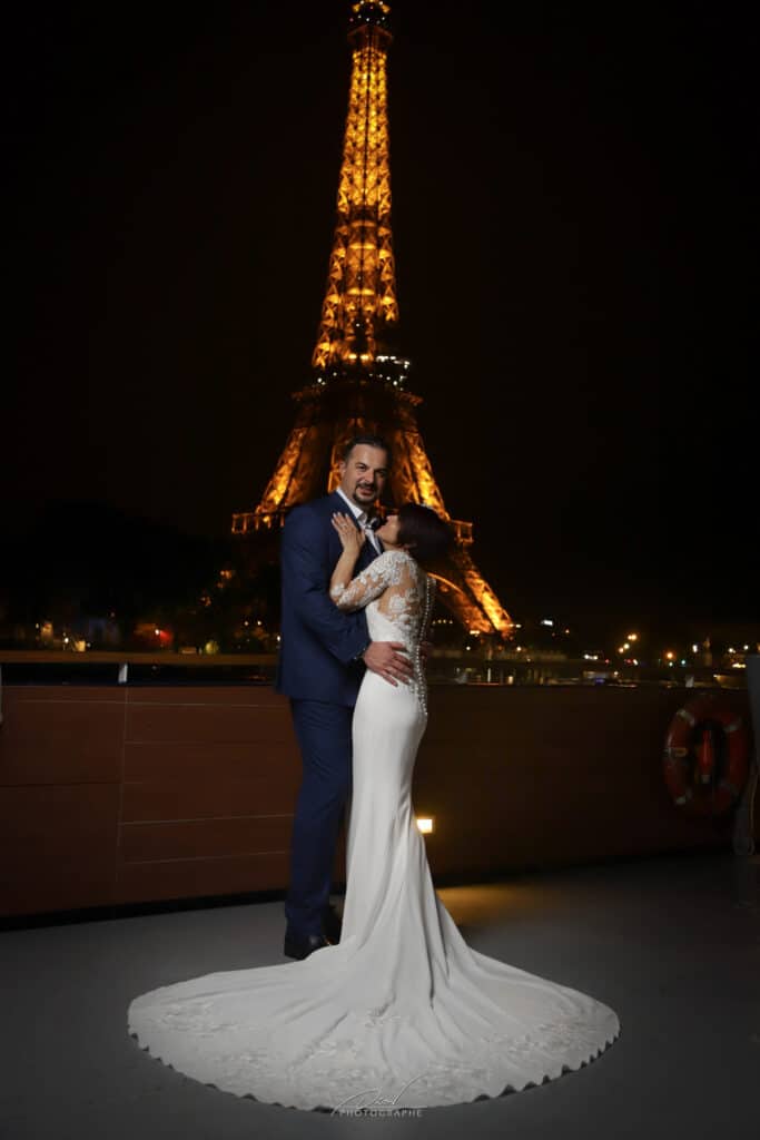 Mariage à Paris: Couple romantique devant la Tour Eiffel Couple marié s'embrassant la nuit devant la Tour Eiffel illuminée à Paris. Robe blanche, costume. "Orion Photographe" visible.