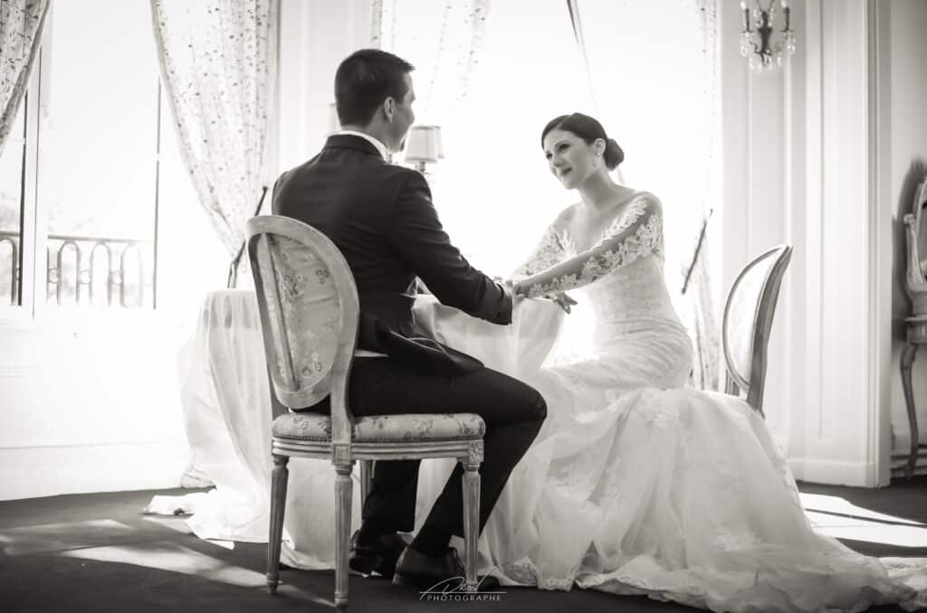 Mariés assis face à face dans un salon lumineux, main dans la main sur une table. La mariée en robe de dentelle, le marié en costume.
