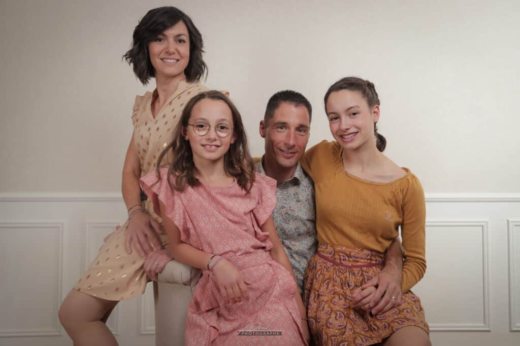 Portrait de famille souriante, mère, père et leurs 2 filles assises sur un canapé. Filigrane Adeline P... PHOTOGRAPHE.