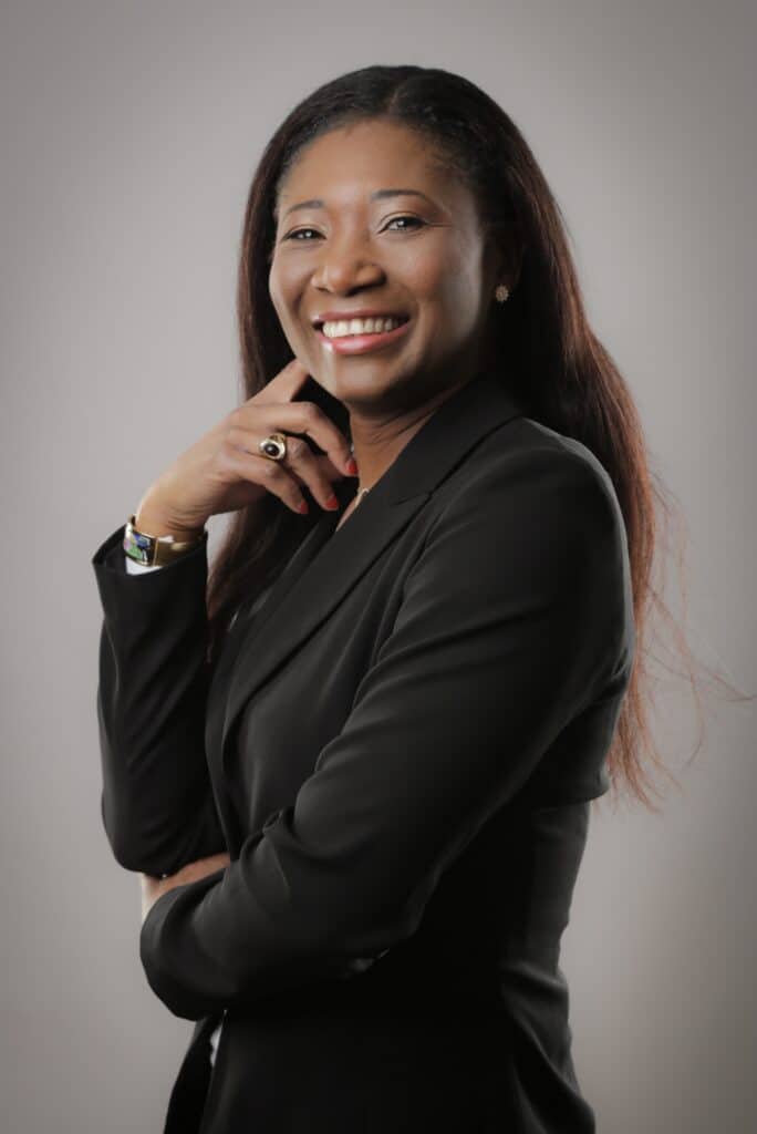 Portrait professionnel femme souriante costume noir Femme souriante en blazer noir, la main près du visage, posant avec confiance sur fond gris.