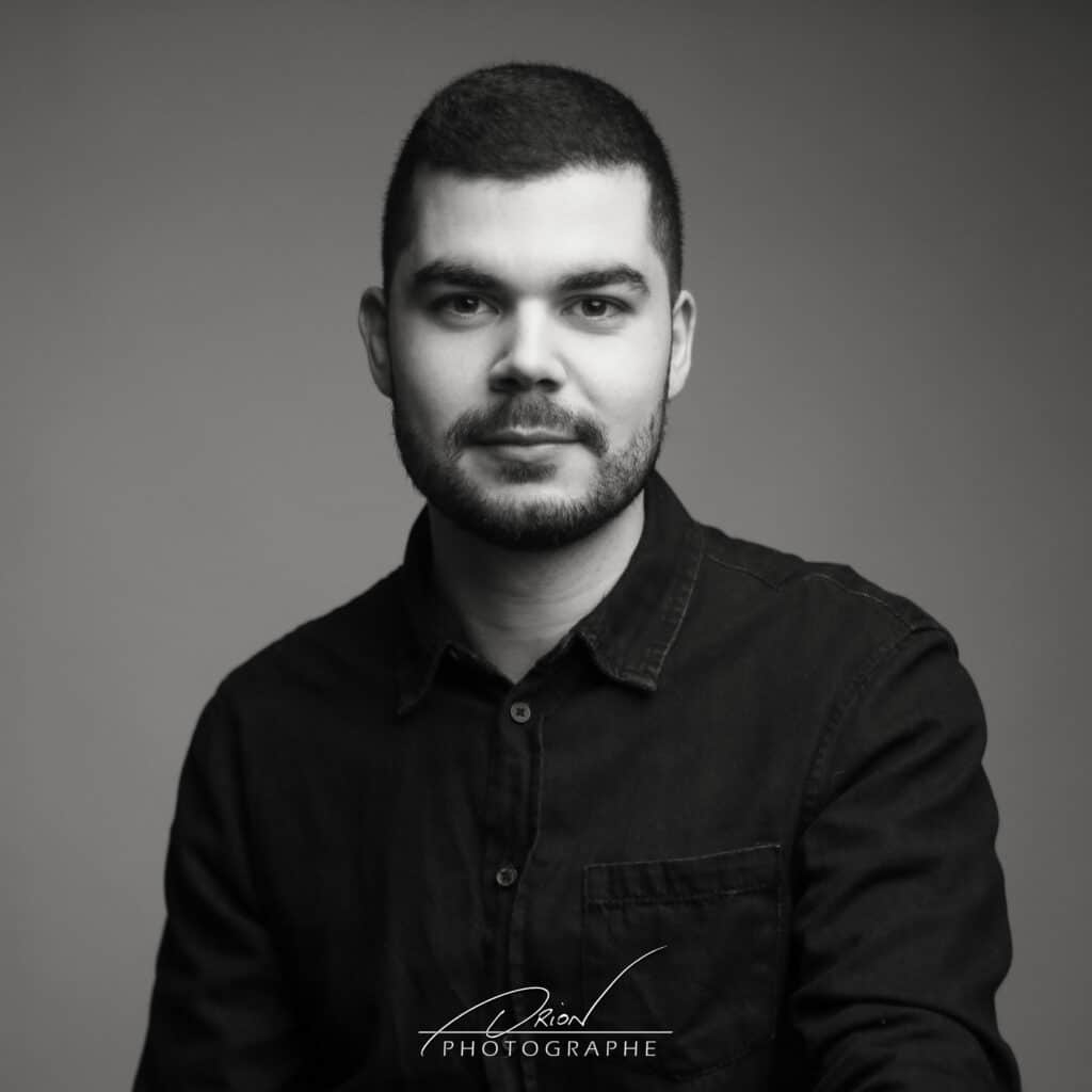 Portrait studio noir et blanc d'un jeune homme barbu en chemise foncée. Regard calme face caméra. Signature Adrion Photographe.